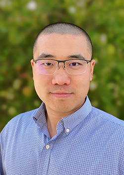 A man with short hair and glasses wearing a blue checkered shirt, standing outdoors with a blurred green background.